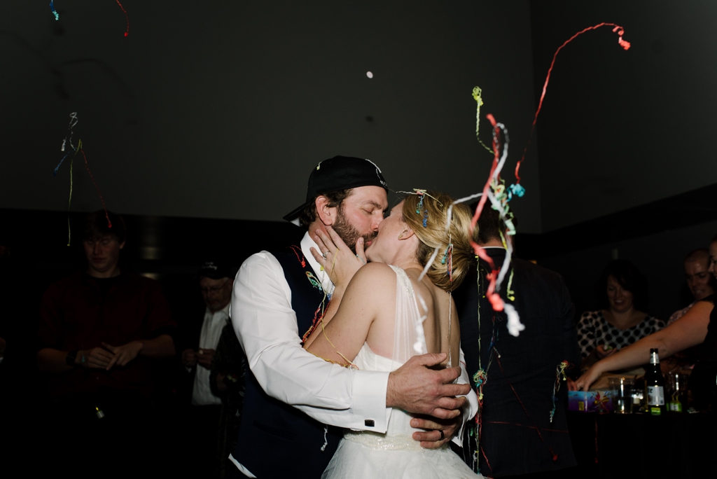 Bride and groom kissing among guests and confetti