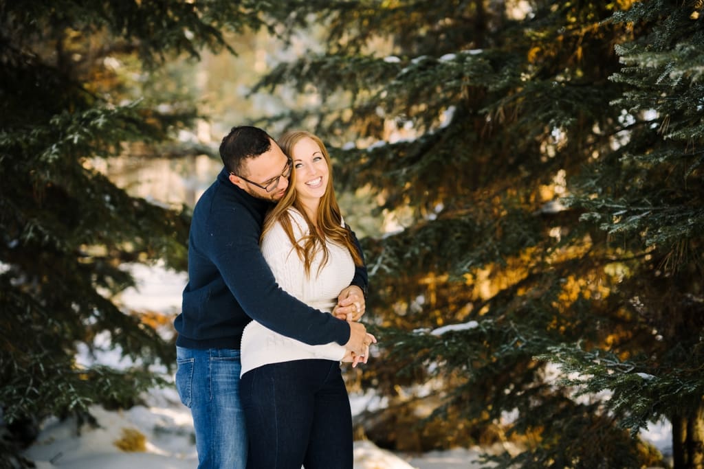 man hugging woman beneath large pine trees at sunset