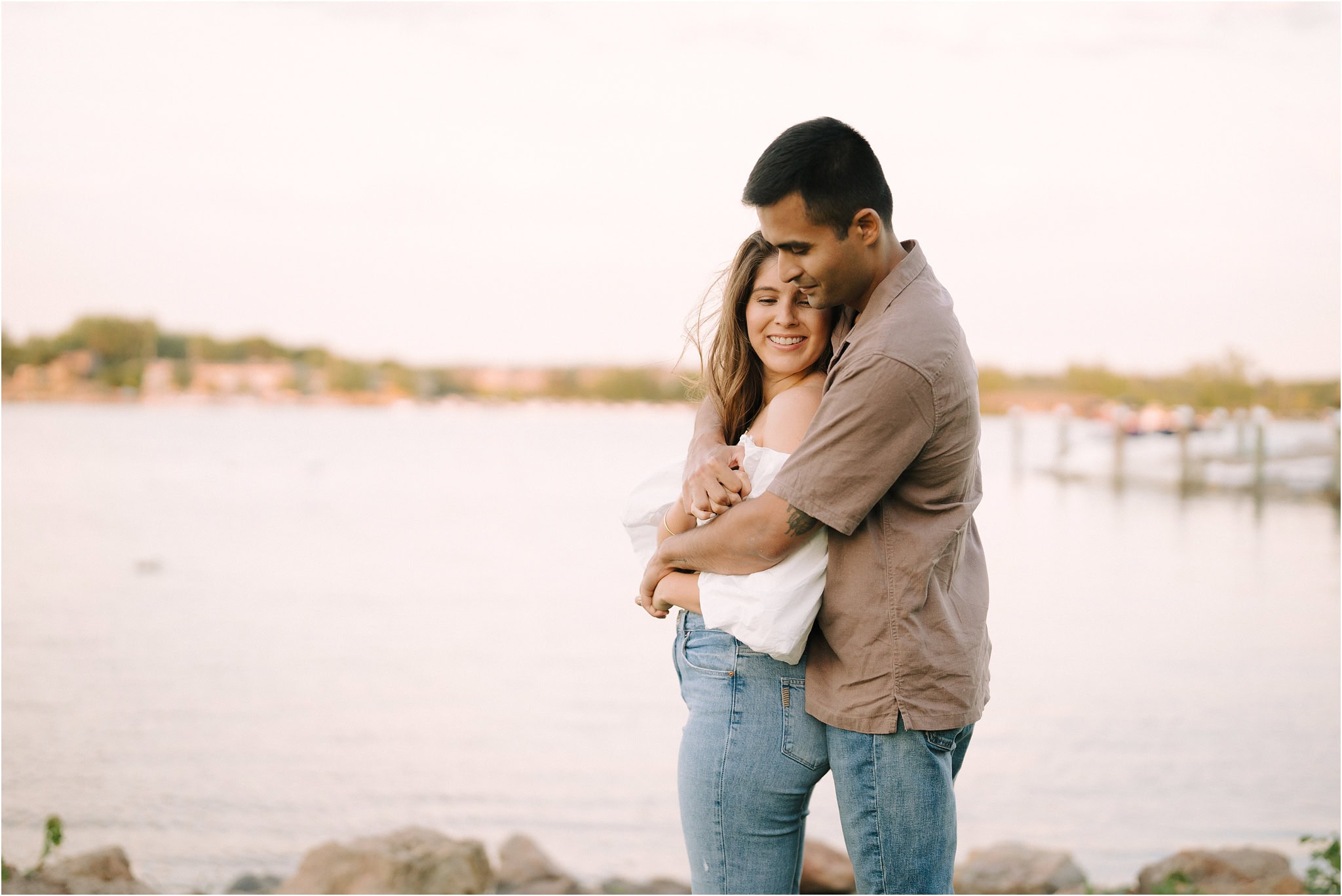 A close-up of a woman smiling shyly while her fiancé hugs her from behind, surrounded by the golden hour light of Port Excelsior.