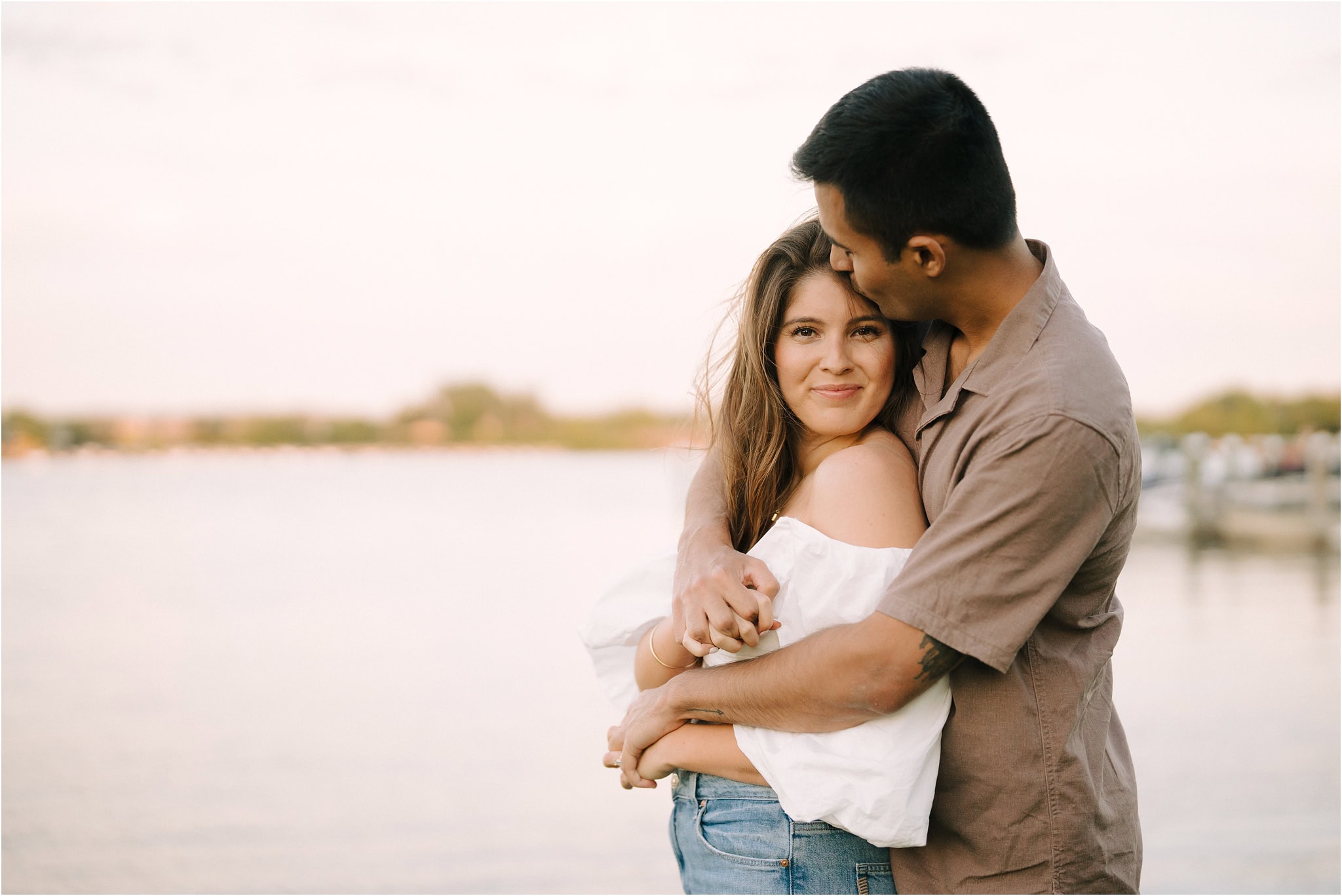 A close-up of a woman smiling at the camera while her fiancé hugs her from behind and kisses the side of her head, surrounded by the golden hour light of Port Excelsior.