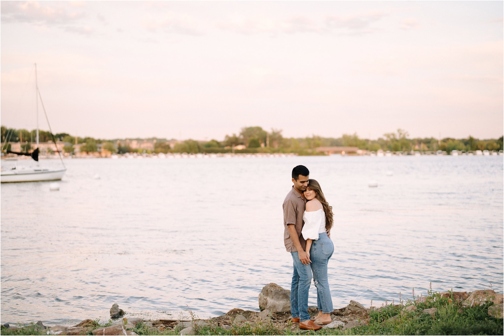 A couple standing before the Port Excelsior lake at golden hour, embracing each other in an intimate moment with her head on his chest.