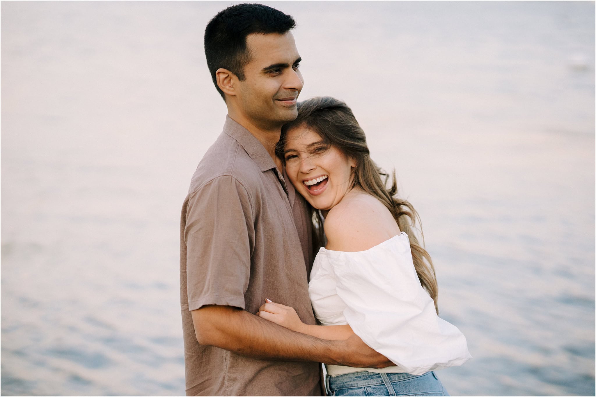 A close-up of a woman laughing with a smile at the camera while her fiancé hugs her, her head on his chest, surrounded by the golden hour light of Port Excelsior.