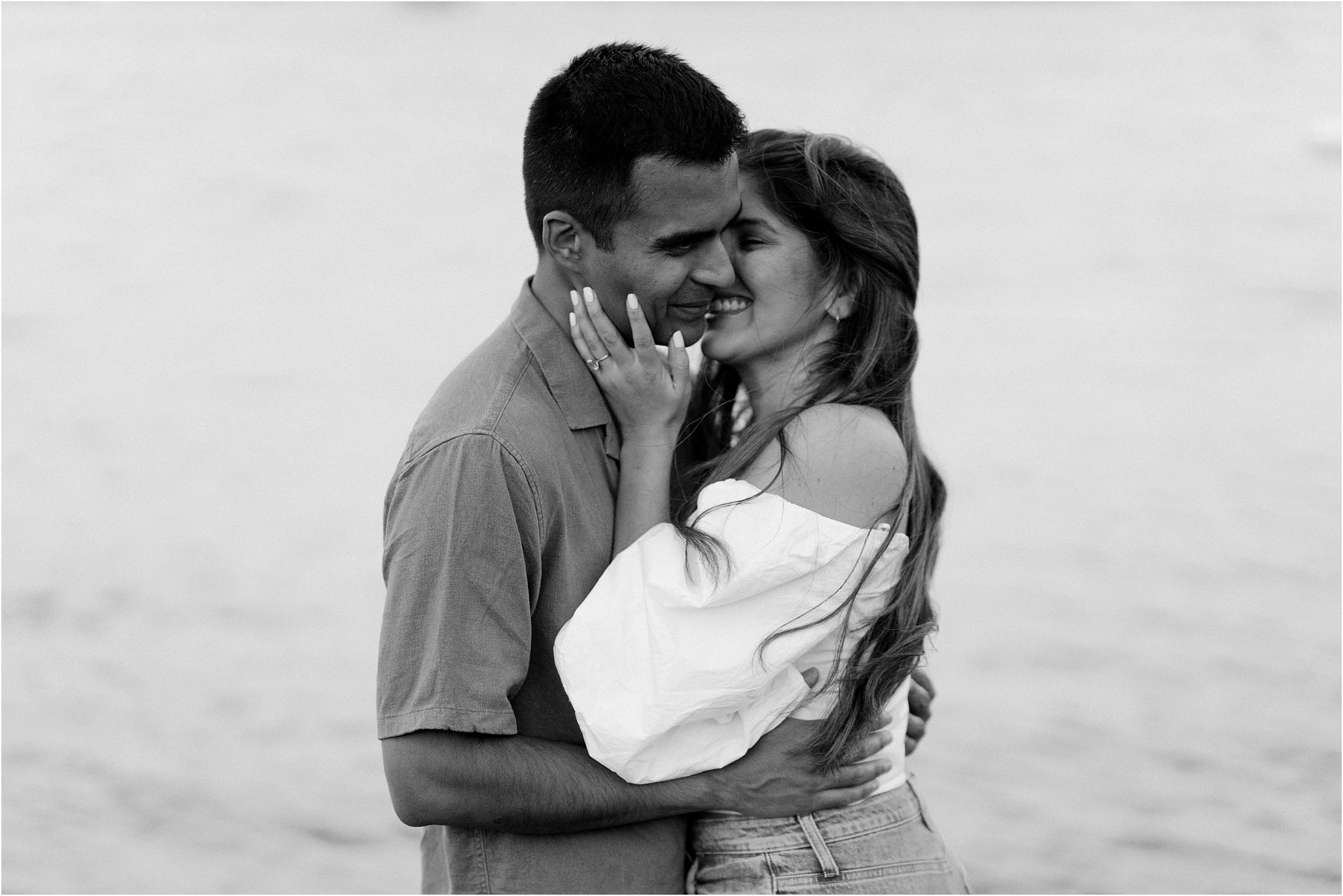 A black and white close-up of an engaged couple embracing, their lips close to kissing as they smile.