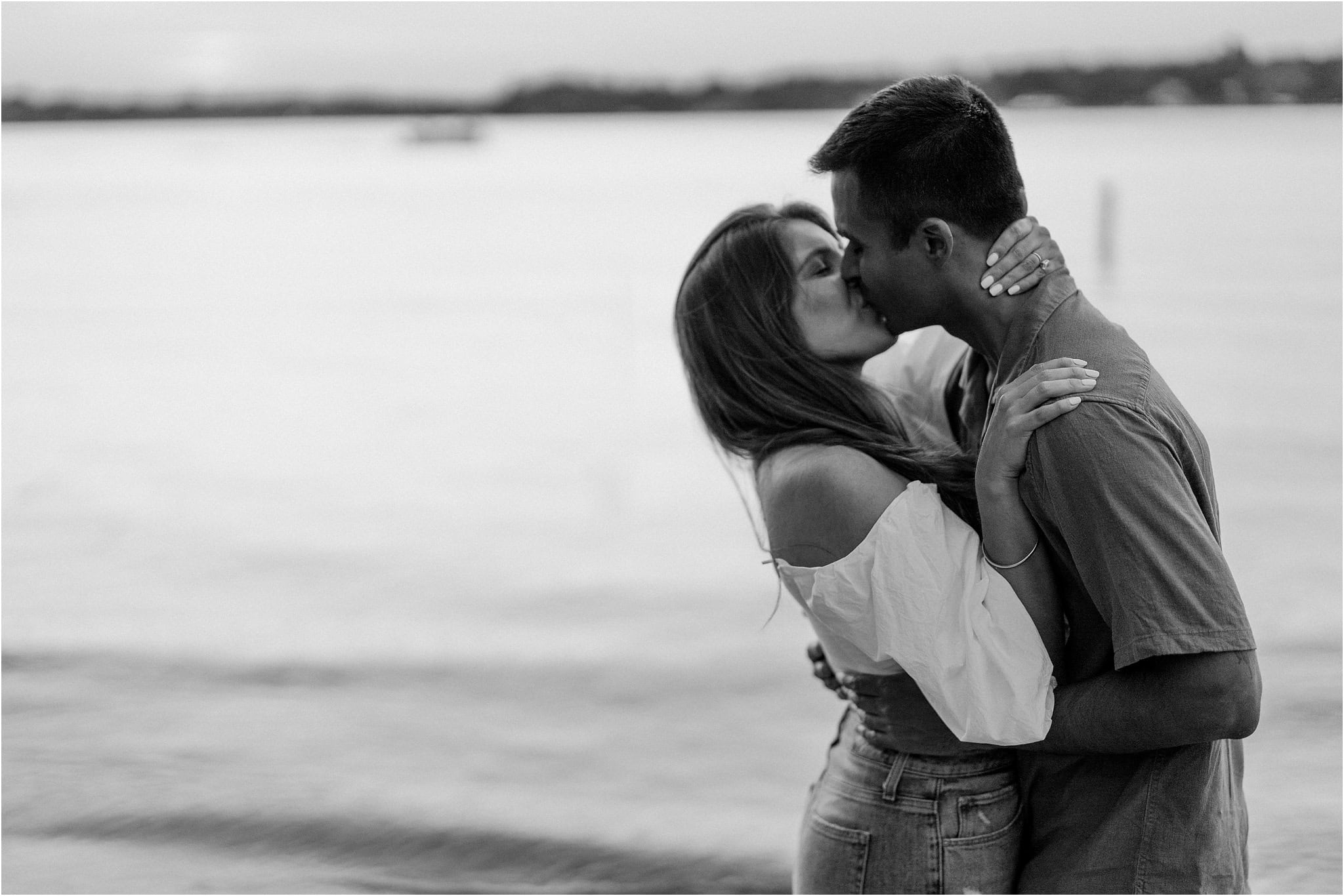 A black and white close-up of an engaged couple embracing in a passionate kiss, the lake at Port Excelsior behind them.