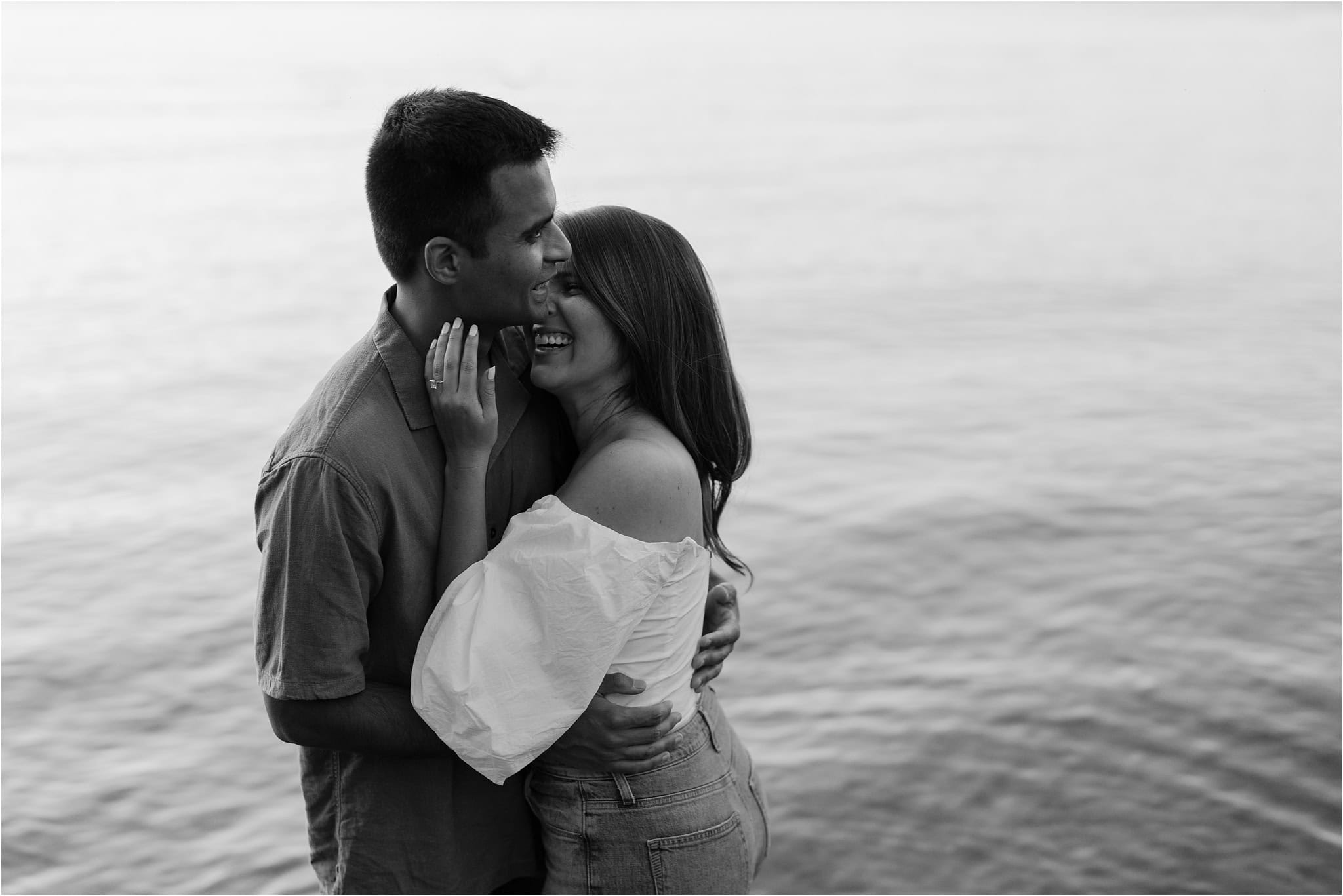 A black and white close-up of an engaged couple laughing with her head on his shoulder and hand held up to his face, the lake at Port Excelsior behind them.