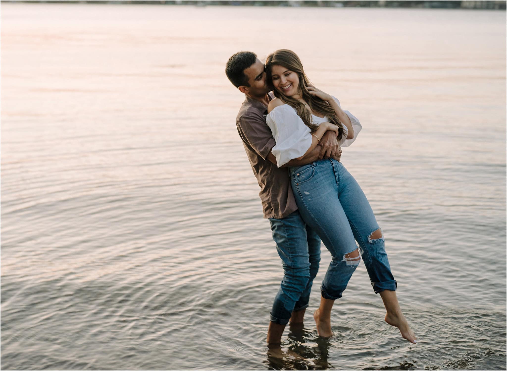 A man hugging his fiancé from behind while lifting her up, kissing her ear as she smiles back at him, ankle deep in the lake at Port Excelsior.